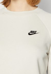 Světlé béžové tričko z měkké látky, s kulatým výstřihem a raglánovými rukávy. Na hrudi je vyšitý černý logo Nike.