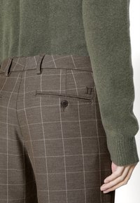 Pantalon à carreaux marron avec poche arrière boutonnée, porté avec un pull en maille à manches longues vert olive, montrant une main reposant le long du corps.