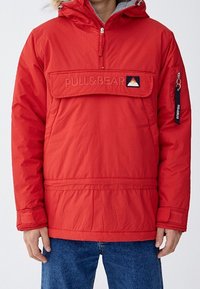 Pullover rosso di Pull&Bear con tasca frontale e cappuccio, indossato da una persona in jeans blu su uno sfondo uniforme.
