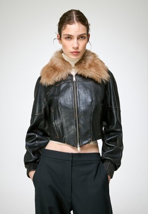 Jeune femme portant une veste en cuir noire courte avec un grand col en fourrure marron, un col roulé blanc et un pantalon noir, debout les mains dans les poches.