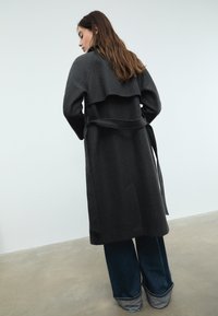 Mörkgrå ullrock med ett lagerdesign, långa ärmar och ett knytbart midjebälte, ihopparat med vida blå denimbyxor.