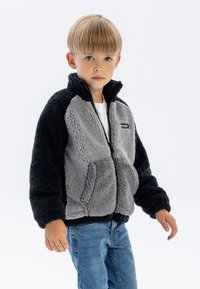 Veste en polaire avec des panneaux gris et noirs, fermeture éclair, col haut et poches latérales. Portée par-dessus une chemise blanche et associée à un jean bleu.