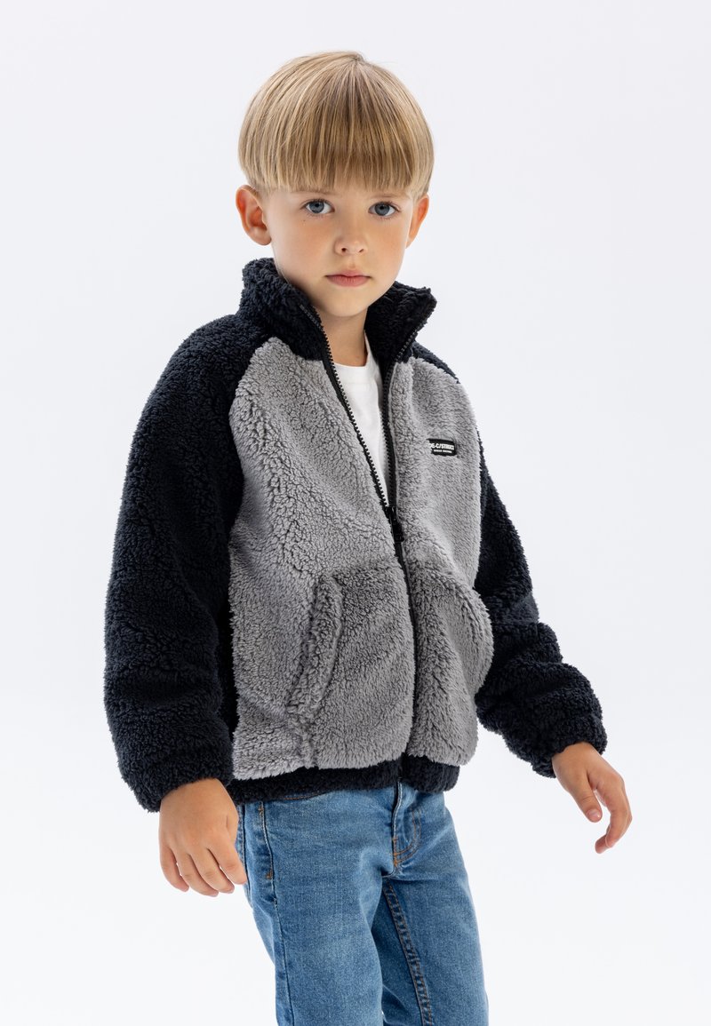 Veste en polaire avec des panneaux gris et noirs, fermeture éclair, col haut et poches latérales. Portée par-dessus une chemise blanche et associée à un jean bleu.