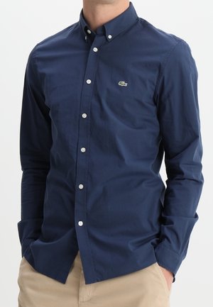 Homme portant une chemise ajustée à manches longues bleu marine avec un petit logo de crocodile vert, associée à un pantalon beige clair.