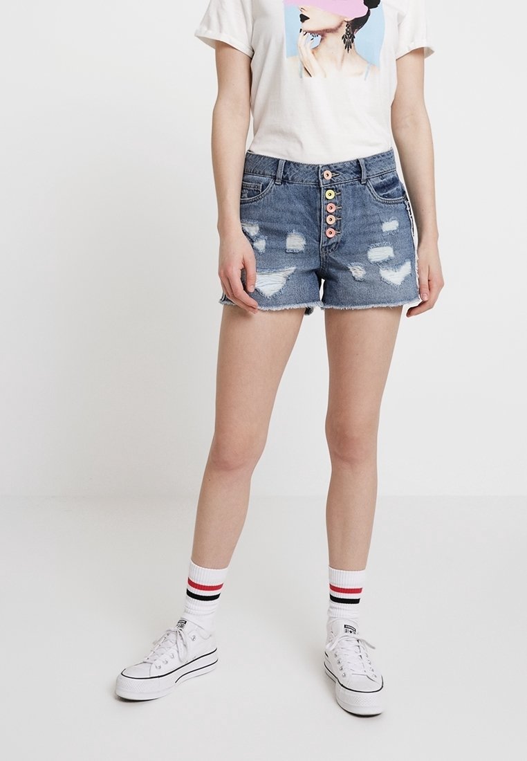 Shorts di jeans, blu chiaro, logori con orlo sfrangiato; caratterizzati da una chiusura a cinque bottoni con bottoni colorati. Abbinati a una t-shirt bianca e sneakers.