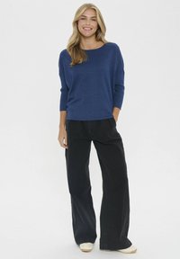 Pulover tricotat de culoare bleumarin, cu un decolteu larg și mâneci trei sferturi, asortat cu pantaloni largi negri cu talie înaltă și încălțăminte deschisă.