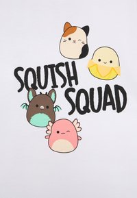 T-shirt blanc avec quatre visages d'animaux en cartoon : un chat calico, un poussin jaune, une chauve-souris brune avec des teintes turquoise, et une créature rose avec des ailes. Le texte indique "SQUISH SQUAD."