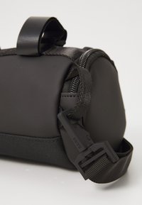 Borsa cilindrica nera con zip, tracolla regolabile e clip in plastica con etichetta "RAINS" su una superficie bianca.