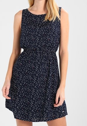 Woman wearing sleeveless navy blue dress with white and red polka dots, elastic waist, and knee-length hem, standing against a plain background.