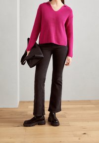 Femme portant un pull rose vif, un jean noir évasé, des bottes noires épaisses, tenant un sac noir plié sur un fond uni.