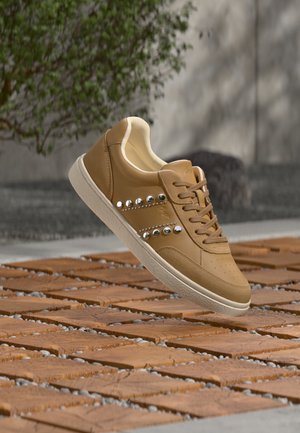 Brown leather sneaker with decorative silver studs on the side, beige lining, and white sole, floating above textured outdoor tiles.