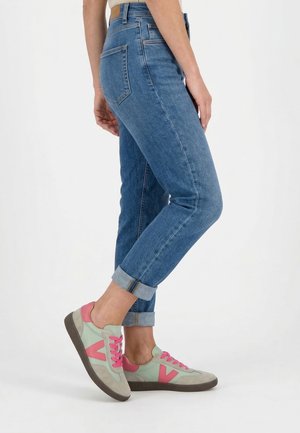 Person iført opkapslede blå jeans og grønne sneakers med lyserøde snørebånd og detaljer, stående mod en hvid baggrund.