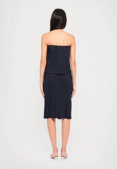 Vrouw in een marineblauwe strapless top met een streepdessin en een bijpassende knie-lange rok, staand met haar rug naar de camera tegen een effen achtergrond.