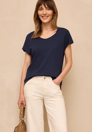 Femme souriante, vêtue d'un haut bleu marine à manches courtes et d’un pantalon crème taille haute, tenant à côté d’elle un sac à franges couleur camel.