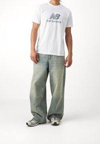 Bílý bavlněný tričko s logem "New Balance", spárované se světle modrými širokými džíny a šedými teniskami s modrými akcenty.