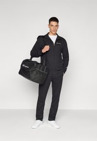Schwarze Zip-Jacke und passende Hose mit einem weißen T-Shirt, hält eine schwarze Sporttasche mit Netz-Akzenten und Logo, vor einem neutralen Hintergrund.