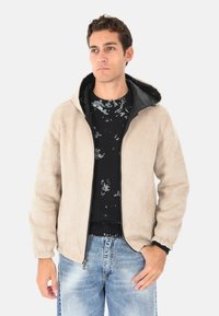 Un giovane uomo indossa una giacca con cappuccio beige sopra un maglione nero a motivo e jeans chiari blu sbiaditi, in piedi con un'espressione neutra.