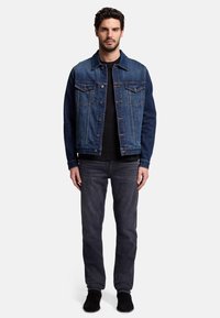Giacca di denim con una finitura blu sbiadita, cinque bottoni, due tasche sul petto e una camicia nera a maniche lunghe sotto; abbinata a jeans scuri e scarpe.