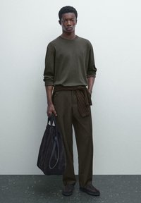 Dunkelgrüner Pullover mit langen Ärmeln, braune Hose mit einem gebundenen Stoffgürtel und einer dunklen Wildleder-Tasche. Das Model steht vor einem hellen Hintergrund.
