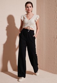 Cremfarbener Crop-Top mit fransigen Ärmeln und elastischem Saum; schwarze weit geschnittene Hose mit seitlichen Taschen; kombiniert mit weißen Sneakern.