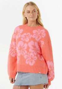 Blonde Frau, die einen leuchtend korallenfarbenen Pullover mit großen pinken Blumenmustern und einem hellblauen Jeans-Minirock trägt.