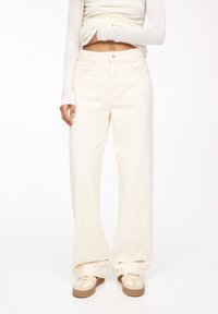 Jean large crème en denim, doté d'un design classique à cinq poches et d'une fermeture à boutons. Associé à des baskets beiges.