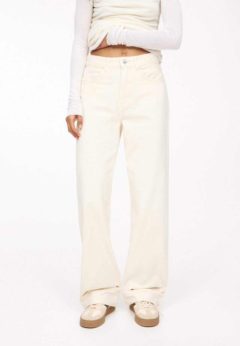 Jean large crème en denim, doté d'un design classique à cinq poches et d'une fermeture à boutons. Associé à des baskets beiges.