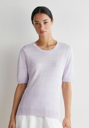 Ung kvinde med mørkt hår iført en lyse lilla kortærmet striktop og hvid nederdel mod en ensfarvet hvid baggrund.