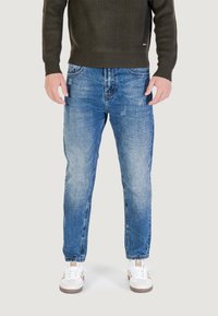 Uomo che indossa jeans blu sbiaditi, sneakers bianche con strisce marroni e un maglione a coste verde oliva scuro, in piedi contro uno sfondo neutro.