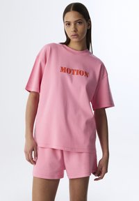 Conjunto de lounge de algodón rosa que incluye una camiseta de corte relajado con "MOTION" en tela contrastante y pantalones cortos a juego. Textura suave, diseño casual.