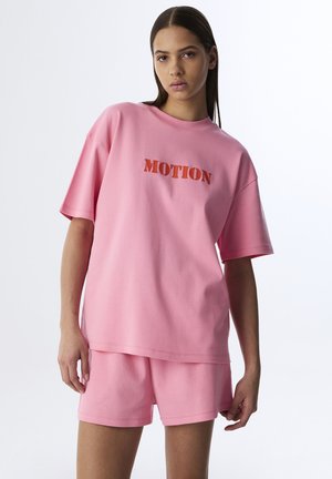 Roze katoenen lounge-set met een relaxed-fit t-shirt waarop "MOTION" staat in vet, contrasterend materiaal en bijpassende shorts. Gladde textuur, casual ontwerp.