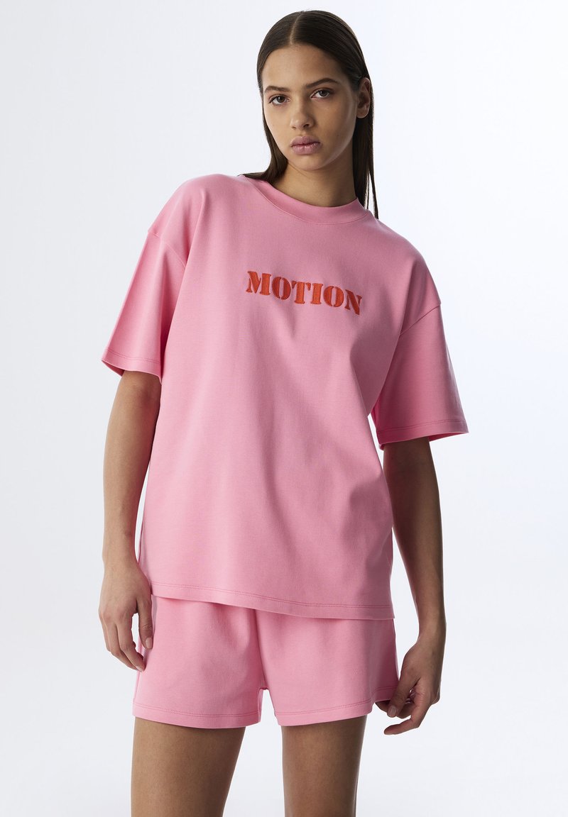 Conjunto de lounge de algodón rosa que incluye una camiseta de corte relajado con "MOTION" en tela contrastante y pantalones cortos a juego. Textura suave, diseño casual.