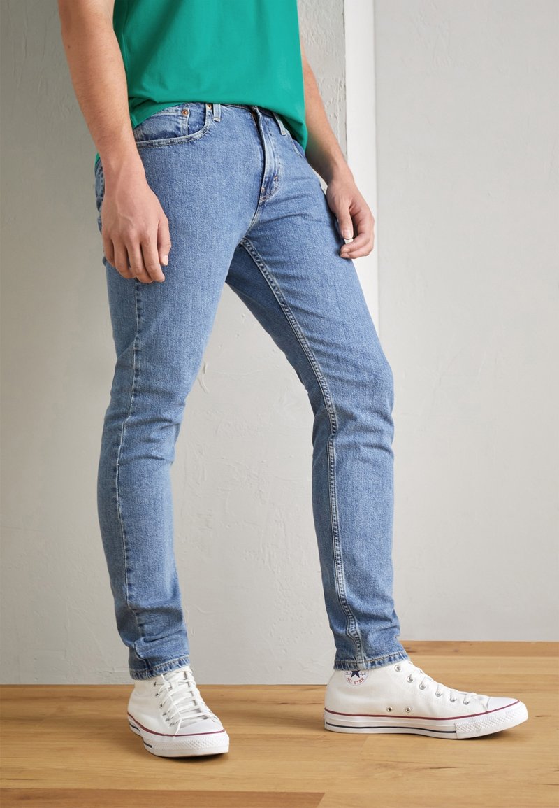 Homme portant un jean slim bleu clair, des baskets blanches montantes et une chemise verte, debout sur un sol en bois devant un mur uni.