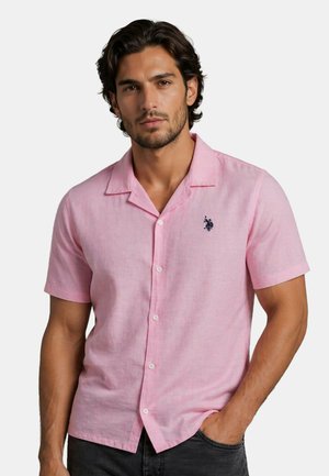 Mann trägt ein hellrosa kurzärmeliges Hemd mit kleinem dunklem gesticktem Logo auf der Brust und posiert mit einer Hand in der Tasche.