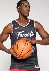 Čierny basketbalový dres s bielymi akcentmi a logom Toronta. Model drží oranžovú basketbalovú loptu, má na sebe bielu čelenku. Hladká textúra látky.