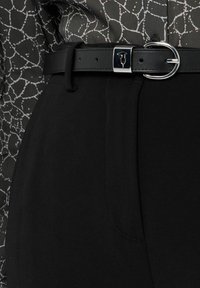 Černé kalhoty z hladké látky s lesklým černým páskem s kovovou přezkou. Košile má geometrický bílý vzor na tmavém podkladu.