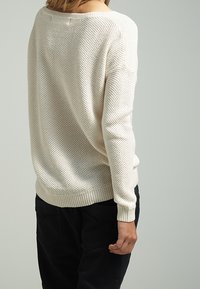 Hellbeige Strickpullover mit lockerer Passform, der ein strukturiertes Muster, gerippte Bündchen und einen runden Halsausschnitt aufweist. Getragen mit dunklen Hosen.