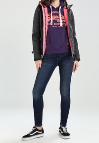 Chaqueta de cremallera negra y rosa sobre un sudadera morada, combinada con jeans oscuros ajustados y zapatillas negras. Texturas suaves, detalles con cordón.