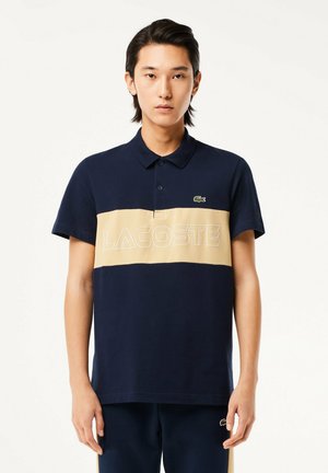 Polo marine avec une rayure horizontale beige comportant "LACOSTE" en lettres blanches. Col rabattable, manches courtes et accent logo.