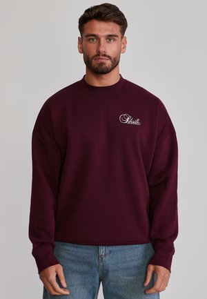 Mann trägt ein bordeauxrotes Sweatshirt mit "Siksilk"-Logo auf der Brust, kombiniert mit blauen Jeans, steht vor einem schlichten grauen Hintergrund.