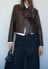 Brązowa skórzana kurtka z krótkim krojem i zapięciem na guzik, zestawiona z ciemnymi spodniami dżinsowymi o szerokich nogawkach oraz czarną torebką. Biała koszula pod spód.