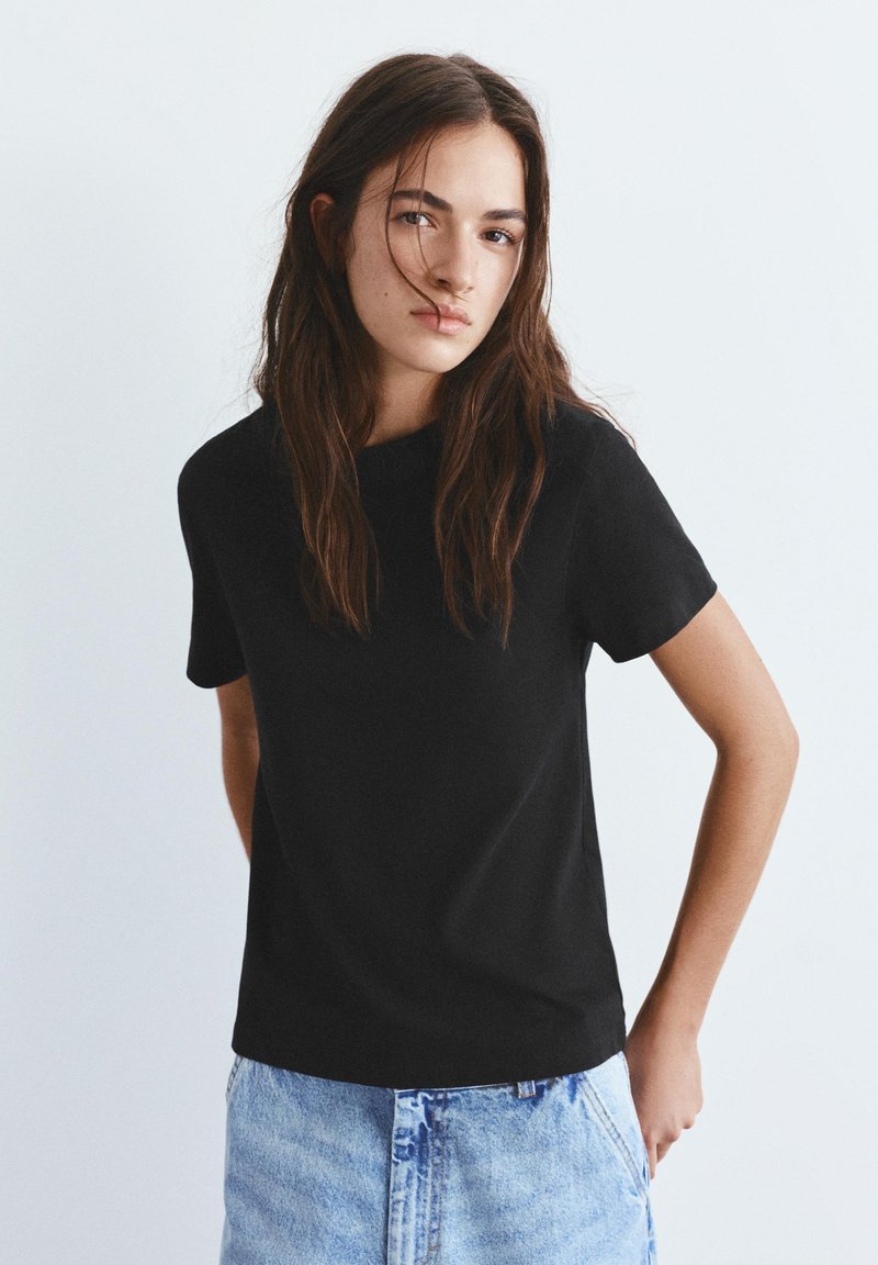 Jeune femme aux longs cheveux bruns portant un t-shirt noir à manches courtes et une jupe en denim bleu clair, debout devant un fond uni clair.