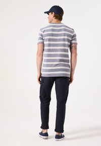 Gestreepte T-shirt met korte mouwen in grijs en marineblauw, gecombineerd met marineblauwe cargobroeken. Model draagt een marineblauwe pet en blauwe sneakers met witte accenten.