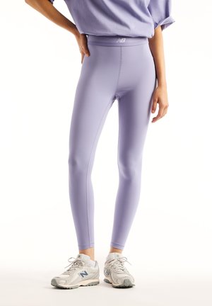 Kvinde iført lyse lilla højtaljede leggings og matchende top, stående med den ene hånd på hoften, iført hvide New Balance sneakers.