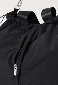 Schwarzer Zugbeutel aus glattem Stoff mit einem Reißverschlussfach und breiten Trägern. Verfügt über einen gerafften oberen Teil und minimalistische Hardwaredetails.