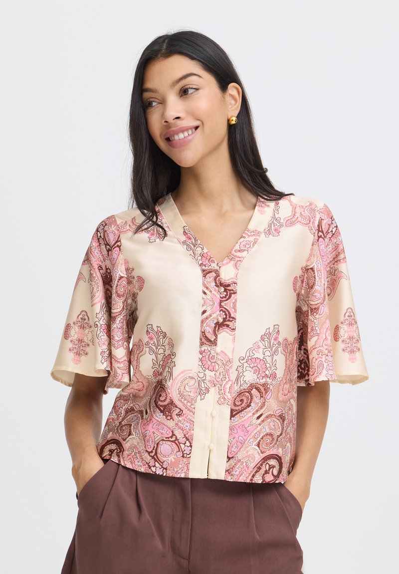 Femme portant un chemisier à motifs crème et rose avec des manches courtes volantées et un pantalon marron, souriante et regardant sur le côté.