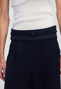 Granatowe spodnie z teksturowanym pasem, z warstwowym materiałem i okrąłym metalowym akcentem, w połączeniu z jasnobeżowym topem bez rękawów.