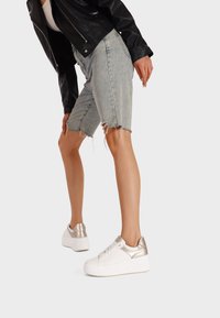 Sneaker platform bianchi con accenti metallici argento, lati testurizzati e design con lacci. Indossati con shorts di denim strappati e una giacca nera.