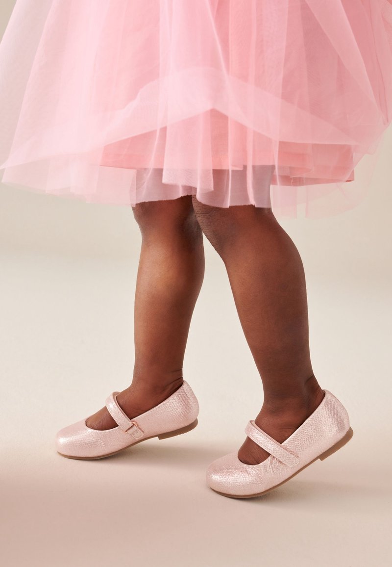 Enfant portant une robe en tulle rose et des chaussures Mary Jane roses brillantes, debout sur un fond neutre.