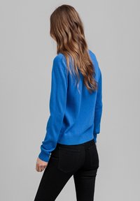 Niebieski sweter z okrągłym dekoltem i długimi rękawami, wykonany z miękkiego materiału. W połączeniu z czarnymi dżinsami; casualowy fason widziany od tyłu.